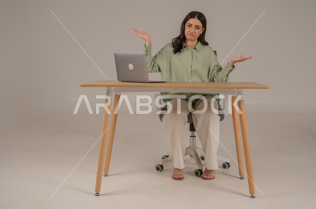 Hesitation and lack of knowledge about something, feeling confused and ignorant about something, using modern and advanced technologies, portrait of a Saudi Arabian Gulf woman wearing casual clothes sitting at an office table raising her hands with gestures of uncertainty, full body image, gray background
