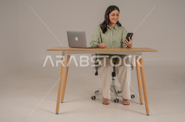 مهن ووظائف مكتبة، انجاز العمل عن بعد، استخدام التقنيات الحديثة والمتطورة، بورتريه لامراة عربية خليجية سعودية ترتدي الزي الكاجوال تجلس على طاولة المكتب تحمل بيدها الهاتف المحمول، صورة لكامل الجسم، خلفية رمادية