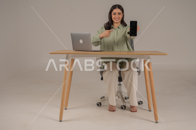 عرض شاشة سوداء فارغة، مهن ووظائف مكتبة، انجاز العمل عن بعد، بورتريه لامراة عربية خليجية سعودية ترتدي الزي الكاجوال تجلس على طاولة المكتب تحمل بيدها الهاتف المحمول، صورة لكامل الجسم، خلفية رمادية