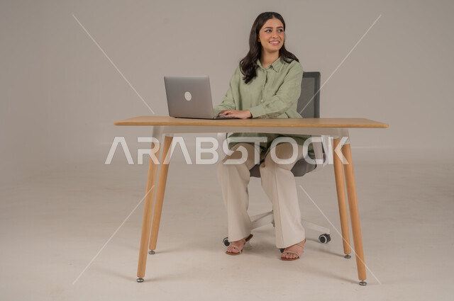 مهن ووظائف مكتبة، انجاز العمل عن بعد عن طريق الانترنت، بورتريه لامراة عربية خليجية سعودية ترتدي الزي الكاجوال تجلس على طاولة المكتب تعمل على جهاز الحاسوب المحمول، صورة لكامل الجسم، خلفية رمادية