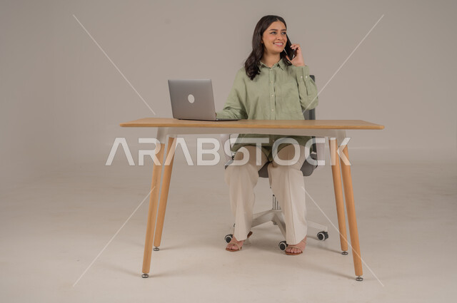 اجراء مكالمات هاتفية، مهن ووظائف مكتبة، استخدام التقنيات الحديثة والمتطورة، انجاز مهام العمل عن بعد، بورتريه من الجانب لامراة عربية خليجية سعودية ترتدي الزي الكاجوال تجلس على طاولة المكتب تحمل بيدها الهاتف المحمول، صورة لكامل الجسم، خلفية رمادية