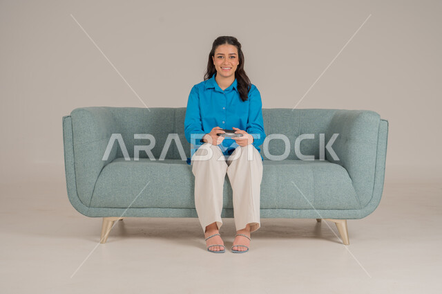 الاستمتاع بألعاب الفيديو في المنزل، استخدام التقنيات التكنولوجية الحديثة، بورتريه لشابة عربية خليجية سعودية ترتدي الزي الكاجوال تجلس على كنبة مريحة تمسك بيدها عصا التحكم بإيماءات الحماس، الأناقة والاهتمام بالمظهر الخارجي، الجسم بالطول الكامل، خلفية رمادية