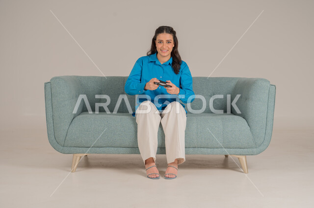 الاستمتاع بألعاب الفيديو في المنزل، استخدام التقنيات التكنولوجية الحديثة، بورتريه لشابة عربية خليجية سعودية ترتدي الزي الكاجوال تجلس على كنبة مريحة تمسك بيدها عصا التحكم بإيماءات الحماس، الأناقة والاهتمام بالمظهر الخارجي، الجسم بالطول الكامل، خلفية رمادية