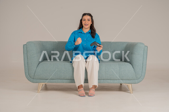 الاستمتاع بألعاب الفيديو في المنزل، استخدام التقنيات التكنولوجية الحديثة، بورتريه لشابة عربية خليجية سعودية ترتدي الزي الكاجوال تجلس على كنبة مريحة تمسك بيدها عصا التحكم بإيماءات الحماس، الأناقة والاهتمام بالمظهر الخارجي، الجسم بالطول الكامل، خلفية رمادية