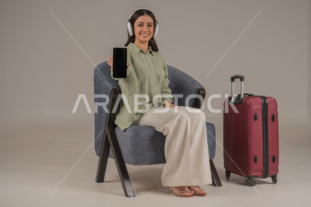عرض شاشة سوداء فارغة، استخدام التكنولوجيا و الأجهزة التقنية الحديثة المتطورة، بورتريه لامراة عربية خليجية سعودية ترتدي الزي الكاجوال وسماعة الهيدفون تجلس على اريكة مريحة بجانب حقيبة سفر تحمل بيدها الهاتف المحمول، صورة لكامل الجسم، خلفية رمادية