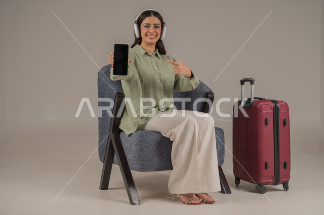 Using advanced modern technology and technical devices, displaying a blank black screen, portrait of a Saudi Arabian Gulf woman wearing casual clothes and headphones sitting on a comfortable sofa next to a travel bag pointing with her index finger to the mobile phone, full body image, gray background