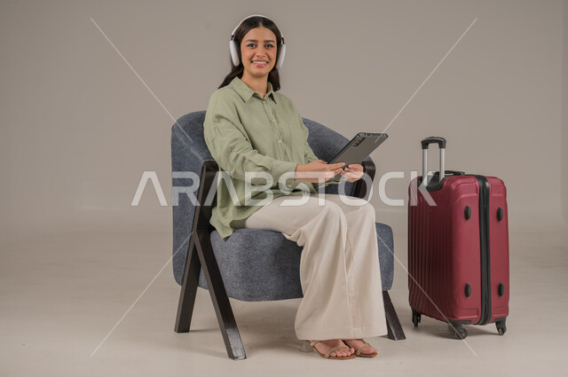 دمج التكنولوجيا المتطورة في الحياة اليومية، استخدام الاجهزة التقنية الحديثة، بورتريه لامراة عربية خليجية سعودية ترتدي الزي الكاجوال وسماعة الهيدفون تجلس على اريكة مريحة بجانب حقيبة سفر تحمل بيدها جهاز التابلت، صورة لكامل الجسم، خلفية رمادية