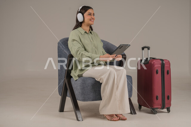 استخدام الاجهزة التقنية الحديثة، دمج التكنولوجيا المتطورة في الحياة اليومية، بورتريه لامراة عربية خليجية سعودية ترتدي الزي الكاجوال وسماعة الهيدفون تجلس على اريكة مريحة بجانب حقيبة سفر تحمل بيدها جهاز التابلت، صورة لكامل الجسم، خلفية رمادية