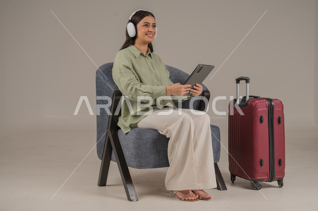 دمج التكنولوجيا المتطورة في الحياة اليومية، استخدام الاجهزة التقنية الحديثة، بورتريه لامراة عربية خليجية سعودية ترتدي الزي الكاجوال وسماعة الهيدفون تجلس على اريكة مريحة بجانب حقيبة سفر تحمل بيدها جهاز التابلت، صورة لكامل الجسم، خلفية رمادية