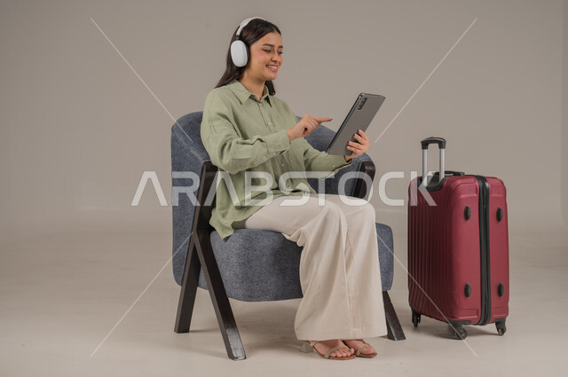 استخدام الاجهزة التقنية الحديثة، دمج التكنولوجيا المتطورة في الحياة اليومية، بورتريه لامراة عربية خليجية سعودية ترتدي الزي الكاجوال وسماعة الهيدفون تجلس على اريكة مريحة بجانب حقيبة سفر تحمل بيدها جهاز التابلت، صورة لكامل الجسم، خلفية رمادية