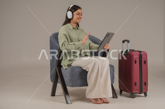 Making a video call, using modern technical devices, integrating advanced technology into daily life, portrait of a Saudi Arabian Gulf woman wearing casual clothes and headphones sitting on a comfortable sofa next to a travel bag holding a tablet in her hand, full body image, gray background
