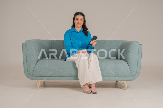دمج التقنيات التكنولوجية الحديثة في الحياة اليومية، بورتريه لشابة عربية خليجية سعودية ترتدي الزي الكاجوال تجلس على كنبة مريحة تمسك بيدها الهاتف المحمول بإيماءات الاندماج والسعادة، تصفح مواقع الانترنت، الاهتمام بالمظهر الخارجي، الجسم بالطول الكامل، خلفية رمادية