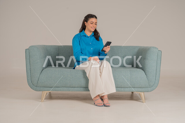 دمج التقنيات التكنولوجية الحديثة في الحياة اليومية، بورتريه لشابة عربية خليجية سعودية ترتدي الزي الكاجوال تجلس على كنبة مريحة تمسك بيدها الهاتف المحمول بإيماءات الاندماج والسعادة، تصفح مواقع الانترنت، الاهتمام بالمظهر الخارجي، الجسم بالطول الكامل، خلفية رمادية