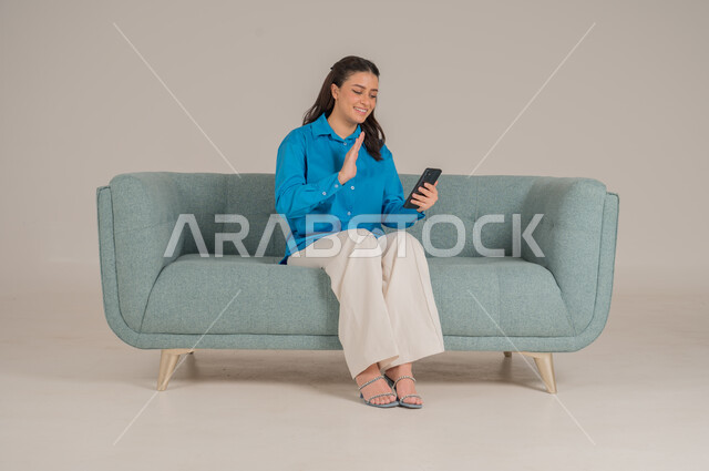 إجراء مكالمات الفيديو بإيماءات الاندماج والسرور، الاستفادة من التقنيات الحديثة في الحياة اليومية، بورتريه لشابة عربية خليجية سعودية ترتدي الزي الكاجوال تجلس على كنبة مريحة تمسك بيدها جهاز الجوال وتتواصل مع الأهل والأصدقاء، الجسم بالطول الكامل، خلفية رمادية