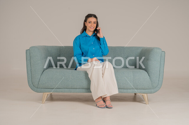  الاستفادة من التقنيات التكنولوجية الحديثة في الحياة اليومية، بورتريه لشابة عربية خليجية سعودية ترتدي الزي الكاجوال تجلس على كنبة مريحة تمسك بيدها الهاتف المحمول وتجري مكالمة هاتفية، التواصل مع الأهل والأصدقاء، الجسم بالطول الكامل، خلفية رمادية