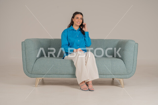 الاستفادة من التقنيات التكنولوجية الحديثة في الحياة اليومية، بورتريه لشابة عربية خليجية سعودية ترتدي الزي الكاجوال تجلس على كنبة مريحة تمسك بيدها الهاتف المحمول وتجري مكالمة هاتفية، التواصل مع الأهل والأصدقاء، الجسم بالطول الكامل، خلفية رمادية