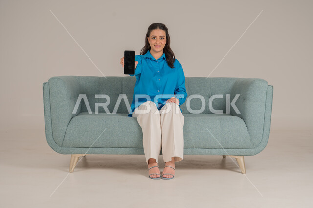 Empty black mobile screen, integration of modern technology into daily life, portrait of a young Saudi Arabian Gulf woman in casual wear sitting on a comfortable sofa holding a mobile phone with gestures of pleasure, concern for appearance, full body, gray background