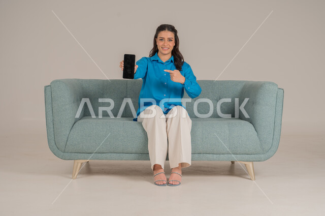 عرض شاشة جوال سوداء فارغة، دمج التقنيات التكنولوجية الحديثة في الحياة اليومية، بورتريه لشابة عربية خليجية سعودية ترتدي الزي الكاجوال تجلس على كنبة مريحة تمسك بيدها الهاتف المحمول بإيماءات السرور، الاهتمام بالمظهر الخارجي، الجسم بالطول الكامل، خلفية رمادية