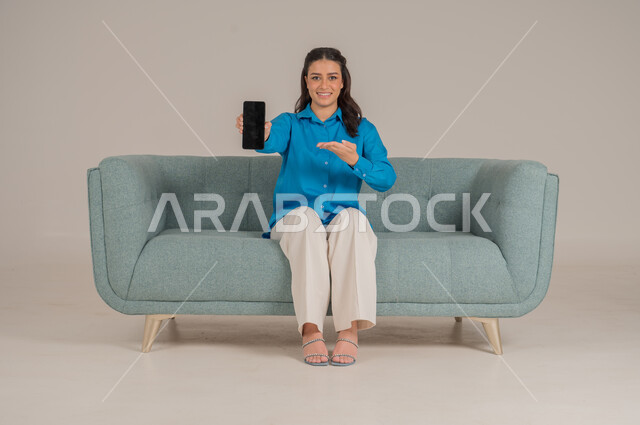 عرض شاشة جوال سوداء فارغة، دمج التقنيات التكنولوجية الحديثة في الحياة اليومية، بورتريه لشابة عربية خليجية سعودية ترتدي الزي الكاجوال تجلس على كنبة مريحة تمسك بيدها الهاتف المحمول بإيماءات السرور، الاهتمام بالمظهر الخارجي، الجسم بالطول الكامل، خلفية رمادية