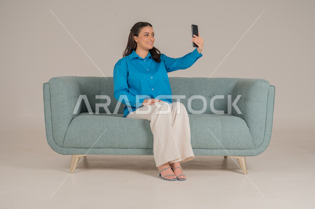 التقاط صور السيلفي التذكارية، استخدام التقنيات الحديثة في الحياة اليومية، بورتريه لشابة عربية خليجية سعودية ترتدي الزي الكاجوال تجلس على كنبة مريحة تمسك بيدها الهاتف المحمول وتقوم بأخذ اللقطات الشخصية بإيماءات السرور، الجسم بالطول الكامل، خلفية رمادية