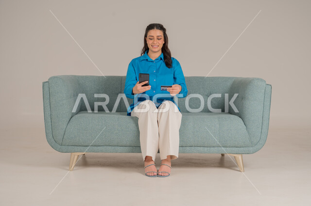 تصفح مواقع الانترنت، دمج التقنيات التكنولوجية الحديثة في الحياة اليومية، بورتريه لشابة عربية خليجية سعودية ترتدي الزي الكاجوال تجلس على كنبة مريحة تمسك بيدها الهاتف المحمول وتشير إليه بإيماءات السرور، الاهتمام بالمظهر الخارجي، الجسم بالطول الكامل، خلفية رمادية