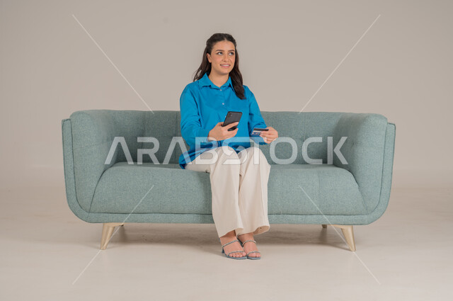 تصفح مواقع الانترنت، دمج التقنيات التكنولوجية الحديثة في الحياة اليومية، بورتريه لشابة عربية خليجية سعودية ترتدي الزي الكاجوال تجلس على كنبة مريحة تمسك بيدها الهاتف المحمول وتشير إليه بإيماءات السرور، الاهتمام بالمظهر الخارجي، الجسم بالطول الكامل، خلفية رمادية