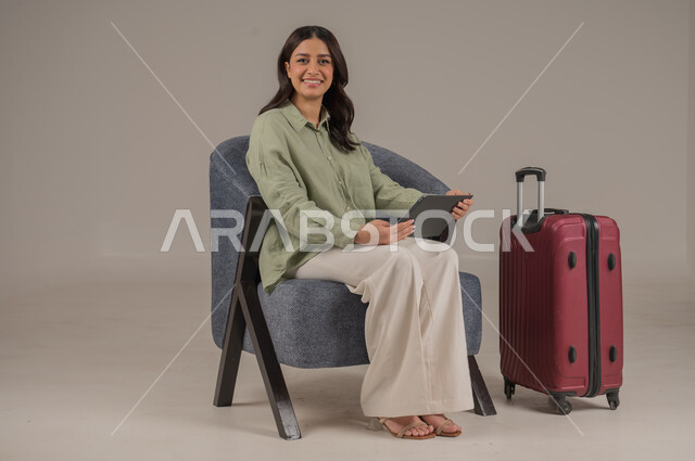 دمج التكنولوجيا المتطورة في الحياة اليومية، الدردشة والتواصل مع الاهل والاصدقاء، استخدام الاجهزة التقنية الحديثة، بورتريه لامراة عربية خليجية سعودية ترتدي الزي الكاجوال تجلس على اريكة مريحة بجانب حقيبة سفر تحمل بيدها جهاز التابلت، صورة لكامل الجسم، خلفية رمادية