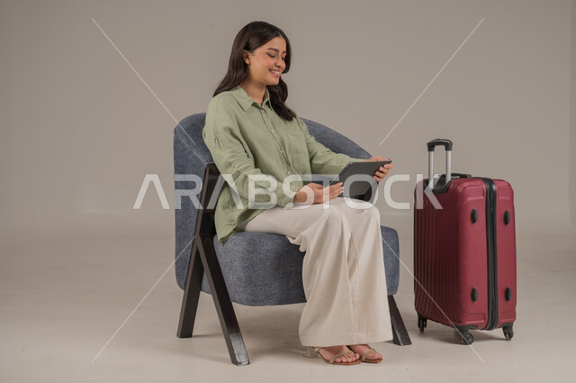 دمج التكنولوجيا المتطورة في الحياة اليومية، الدردشة والتواصل مع الاهل والاصدقاء، استخدام الاجهزة التقنية الحديثة، بورتريه لامراة عربية خليجية سعودية ترتدي الزي الكاجوال تجلس على اريكة مريحة بجانب حقيبة سفر تحمل بيدها جهاز التابلت، صورة لكامل الجسم، خلفية رمادية