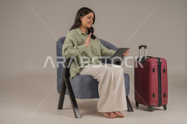 الدردشة والتواصل مع الاهل والاصدقاء، استخدام الاجهزة التقنية الحديثة، دمج التكنولوجيا المتطورة في الحياة اليومية، بورتريه لامراة عربية خليجية سعودية ترتدي الزي الكاجوال تجلس على اريكة مريحة بجانب حقيبة سفر تحمل بيدها جهاز التابلت، صورة لكامل الجسم، خلفية رمادية