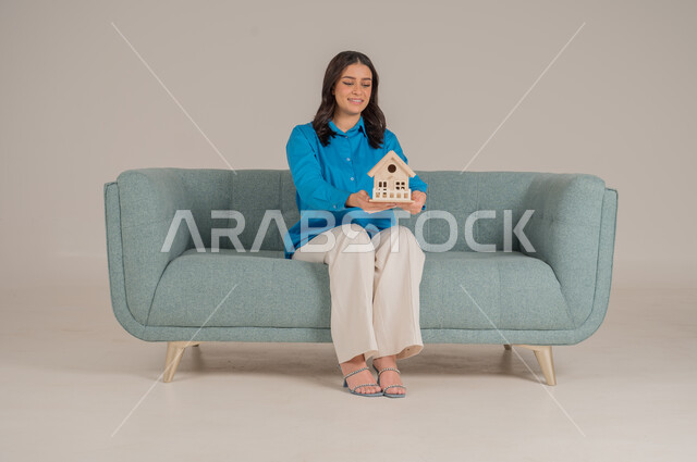 Real estate buying and selling, seeking to own a new home, strong family ties, portrait of a young Saudi Arabian Gulf woman in casual wear sitting on a comfortable sofa holding a small wooden model of a house with gestures of joy and hope, full body, gray background