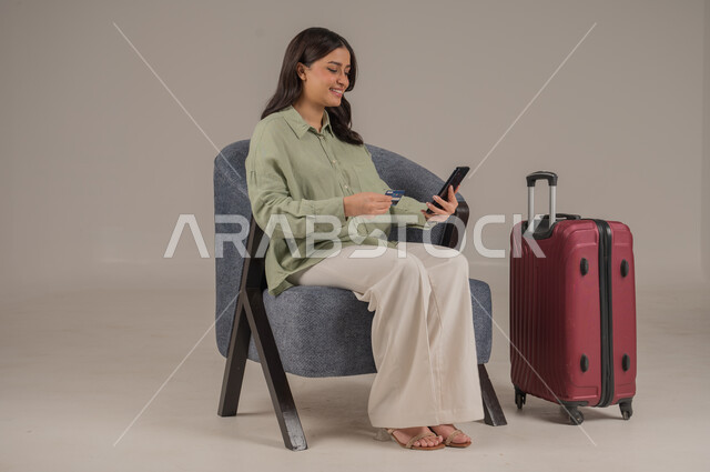 سهولة تسديد الفواتير عبر الحساب البنكي، الشراء والدفع عن طريق بطاقة الصراف الإئتمانية، بورتريه لامراة عربية خليجية سعودية ترتدي الزي الكاجوال تجلس على اريكة مريحة بجانب حقيبة سفر تحمل بيدها الهاتف المحمول وكرت البنك المصرفي، صورة لكامل الجسم، خلفية رمادية