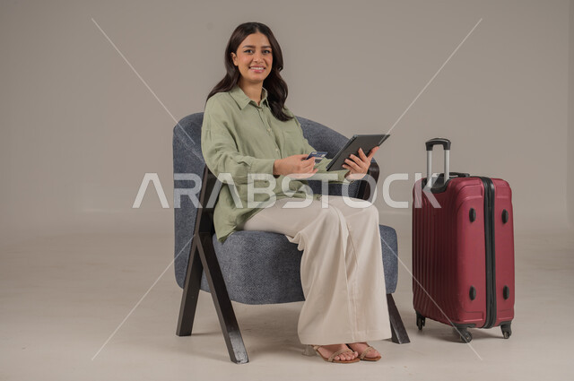 الشراء والدفع عن طريق بطاقة الصراف الإئتمانية، سهولة تسديد الفواتير عبر الحساب البنكي، بورتريه لامراة عربية خليجية سعودية ترتدي الزي الكاجوال تجلس على اريكة مريحة بجانب حقيبة سفر تحمل بيدها التابلت وكرت البنك المصرفي، صورة لكامل الجسم، خلفية رمادية