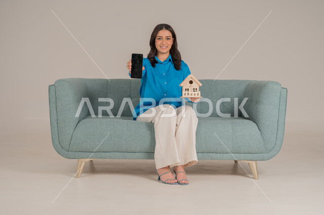 Black blank mobile phone screen, online housing and ownership, online real estate buying and selling, portrait of young Saudi Arabian Gulf woman in casual wear sitting on sofa holding wooden model of house with gestures of happiness and pleasure, full length body, gray background
