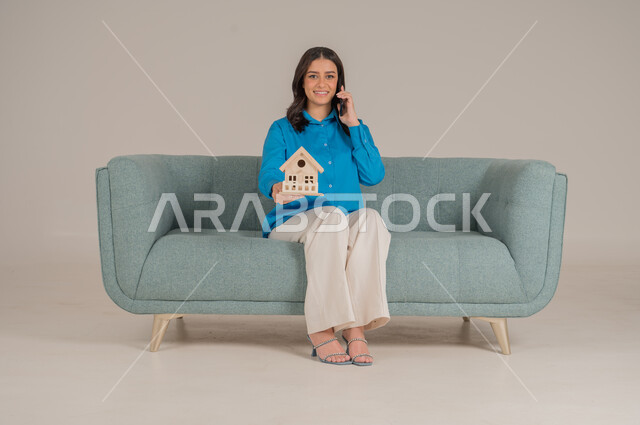 الاستفادة من التقنيات الحديثة في المجال العقاري، الإسكان والتمليك أونلاين عبر الهاتف المحمول، بيع وشراء العقارات عبر الانترنت، بورتريه لشابة عربية خليجية سعودية ترتدي الزي الكاجوال تجلس على كنبة وتحمل بيديها مجسم خشبي لمنزل، الجسم بالطول الكامل، خلفية رمادية