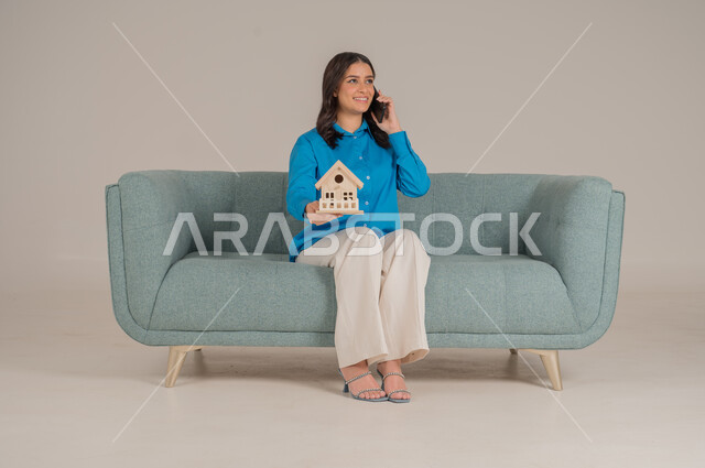 الاستفادة من التقنيات الحديثة في المجال العقاري، الإسكان والتمليك أونلاين عبر الهاتف المحمول، بيع وشراء العقارات عبر الانترنت، بورتريه لشابة عربية خليجية سعودية ترتدي الزي الكاجوال تجلس على كنبة وتحمل بيديها مجسم خشبي لمنزل، الجسم بالطول الكامل، خلفية رمادية