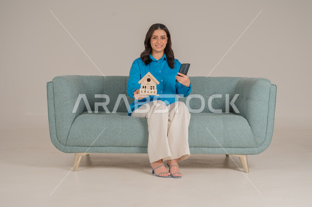 الإسكان والتمليك أونلاين عبر الهاتف المحمول، بيع وشراء العقارات عبر الانترنت، الاستفادة من التقنيات الحديثة في المجال العقاري، بورتريه لشابة عربية خليجية سعودية ترتدي الزي الكاجوال تجلس على كنبة وتحمل بيديها مجسم خشبي لمنزل، الجسم بالطول الكامل، خلفية رمادية