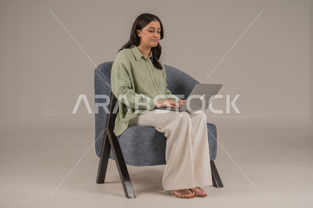 استخدام التقنيات الحديثة والمتطورة، انجاز مهام العمل عن بعد، ايماءات السرور والسعادة، بورتريه لامراة عربية خليجية سعودية ترتدي الزي الكاجوال تجلس على اريكة مريحة تعمل على جهاز الحاسوب المحمول، صورة لكامل الجسم، خلفية رمادية