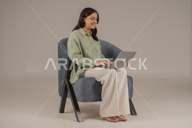 Gestures of pleasure and happiness, using modern and advanced technologies, completing work tasks remotely, portrait of a Saudi Arabian Gulf woman wearing casual clothes sitting on a comfortable sofa working on a laptop, full body image, gray background