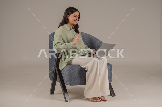 استخدام التقنيات الحديثة والمتطورة، انجاز مهام العمل عن بعد، ايماءات السرور والسعادة، بورتريه لامراة عربية خليجية سعودية ترتدي الزي الكاجوال تجلس على اريكة مريحة تعمل على جهاز الحاسوب المحمول، صورة لكامل الجسم، خلفية رمادية