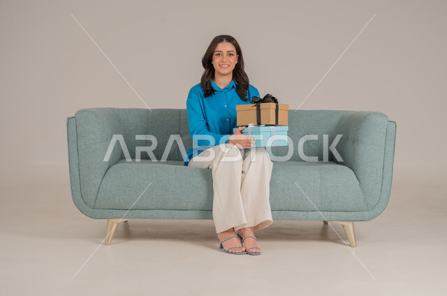The concept of gifting and appreciation, giving gifts on holidays and occasions to express love, portrait of a young Saudi Arabian Gulf woman wearing casual clothes sitting on a sofa holding an elegantly wrapped gift box in her hand and looking at the camera with gestures of pleasure and happiness, full body, gray background