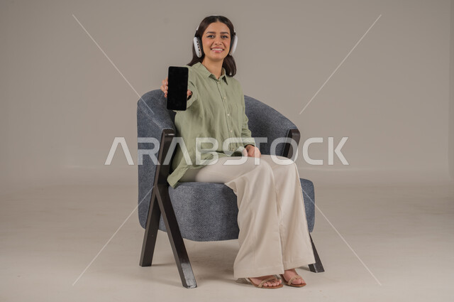 عرض شاشة سوداء فارغة، ايماءات السرور والسعادة، استخدام التقنيات الحديثة والمتطورة، انجاز مهام العمل عن بعد، بورتريه لامراة عربية خليجية سعودية ترتدي الزي الكاجوال وسماعة الهيدفون تجلس على اريكة مريحة تحمل بيدها الهاتف المحمول، صورة لكامل الجسم، خلفية رمادية
