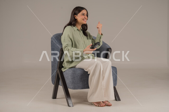 قضاء إجازة صيفية ممتعة، اللعب في ألعاب البلايستيشن الترفيهية، إيماءات الاستمتاع والاندماج والتفاعل، بورتريه لامراة عربية خليجية سعودية ترتدي الزي الكاجوال تجلس على اريكة مريحة تحمل بيدها عصا التحكم، صورة لكامل الجسم، خلفية رمادية