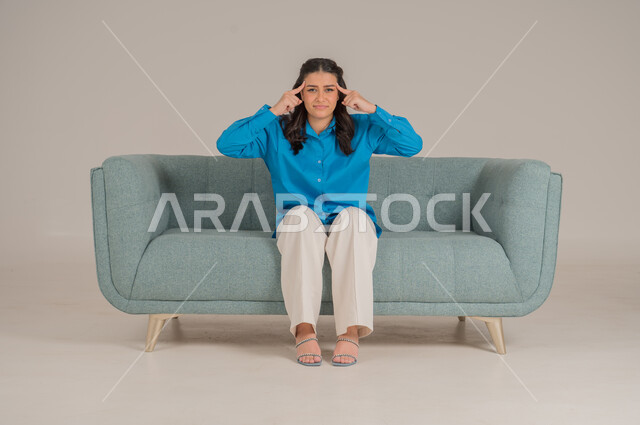 Pointing head with gestures of discomfort and disturbance, feeling pain and headache, portrait of a young Saudi Arabian Gulf woman in casual wear sitting on a comfortable sofa with her index fingers on her head and looking at the camera with gestures of discomfort, full body, gray background