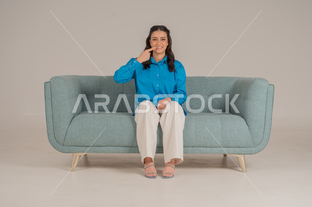 النظر إلى الكاميرا بإيماءات السرور والفرح، أهمية المحافظة على صحة الفم والأسنان من التسوس، بورتريه لشابة عربية خليجية سعودية مبتسمة ترتدي الزي الكاجوال تجلس على كنبة مريحة تشير باصبع السبابة إلى أسنانها، الجسم بالطول الكامل، خلفية رمادية