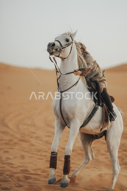  اتباع تقاليد الأجداد برعاية وتربية الجياد، الفروسية وهواية ركوب الأحصنة في الصحراء، أنشطة ترفيهية نسائية، الاهتمام والشغف بركوب الجياد، صورة من الجانب لشابة عربية سعودية خليجية محجبة ترتدي العباءة تقوم بالتجول على ظهر جوادها العربي الأصيل في الصحارى