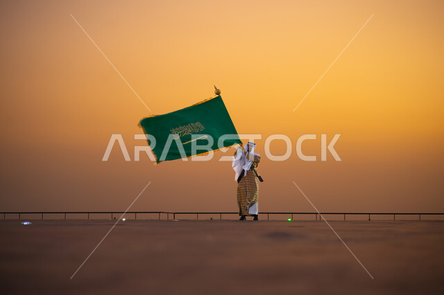 المشاركة بالمناسبات والاعياد الوطنية، يوم العلم 11 مارس, ذكرى تأسيس المملكة 22 فبراير، الاحتفال باليوم الوطني السعودي 23 سبتمبر، صورة من الامام لرجل عربي خليجي سعودي يرتدي زي يوم التأسيس 1727 م ويحمل بيده علم المملكة السعودية وقت غروب الشمس