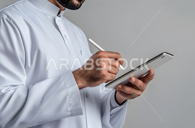 التقنيات الحديثة المتطورة، بورتريه مقرب ليد رجل عربي خليجي سعودي يرتدي الثوب التقليدي يحمل جهاز آيباد ويستخدم قلم ستايلس للكتابة أو الرسم على الشاشة، التسويق لموقع إلكتروني، استخدام قلم التابليت لتدوين الملاحظات، برامج التواصل الاجتماعي، خلفية بيضاء
