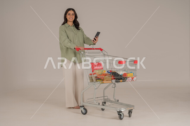 استخدام التقنيات الحديثة, بورتريه امرأة عربية سعودية خليجية ترتدي ثياب كاجوال  تقوم بجر عربة التسوق تحمل بيدها هاتف محمول, قضاء احتياجات المنزل , الإستفادة من العروض والخصومات , الإستمتاع بشراء المستلزمات, صورة الجسم بالطول الكامل, خلفية بيج. 
