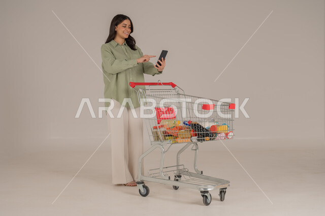 استخدام التقنيات الحديثة, بورتريه امرأة عربية سعودية خليجية ترتدي ثياب كاجوال  تقوم بجر عربة التسوق تحمل بيدها هاتف محمول, قضاء احتياجات المنزل , الإستفادة من العروض والخصومات , الإستمتاع بشراء المستلزمات, صورة الجسم بالطول الكامل, خلفية بيج. 