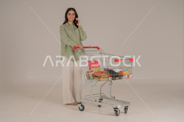 استخدام التقنيات الحديثة, بورتريه امرأة عربية سعودية خليجية ترتدي ثياب كاجوال  تقوم بجر عربة التسوق تحمل بيدها هاتف محمول, قضاء احتياجات المنزل , الإستفادة من العروض والخصومات , الإستمتاع بشراء المستلزمات, صورة الجسم بالطول الكامل, خلفية بيج. 
